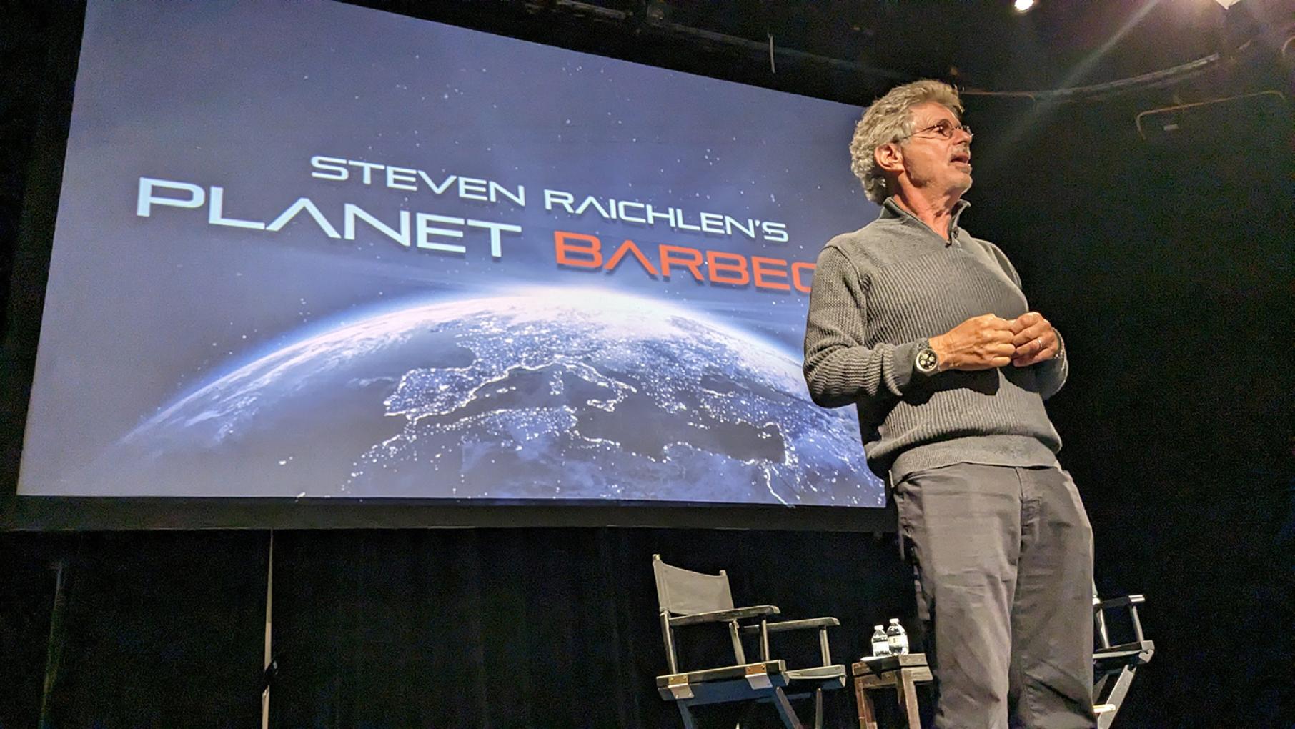 Steven Raichlen stands onstage in front of a screen advertising his Planet Barbecue show at WTTW